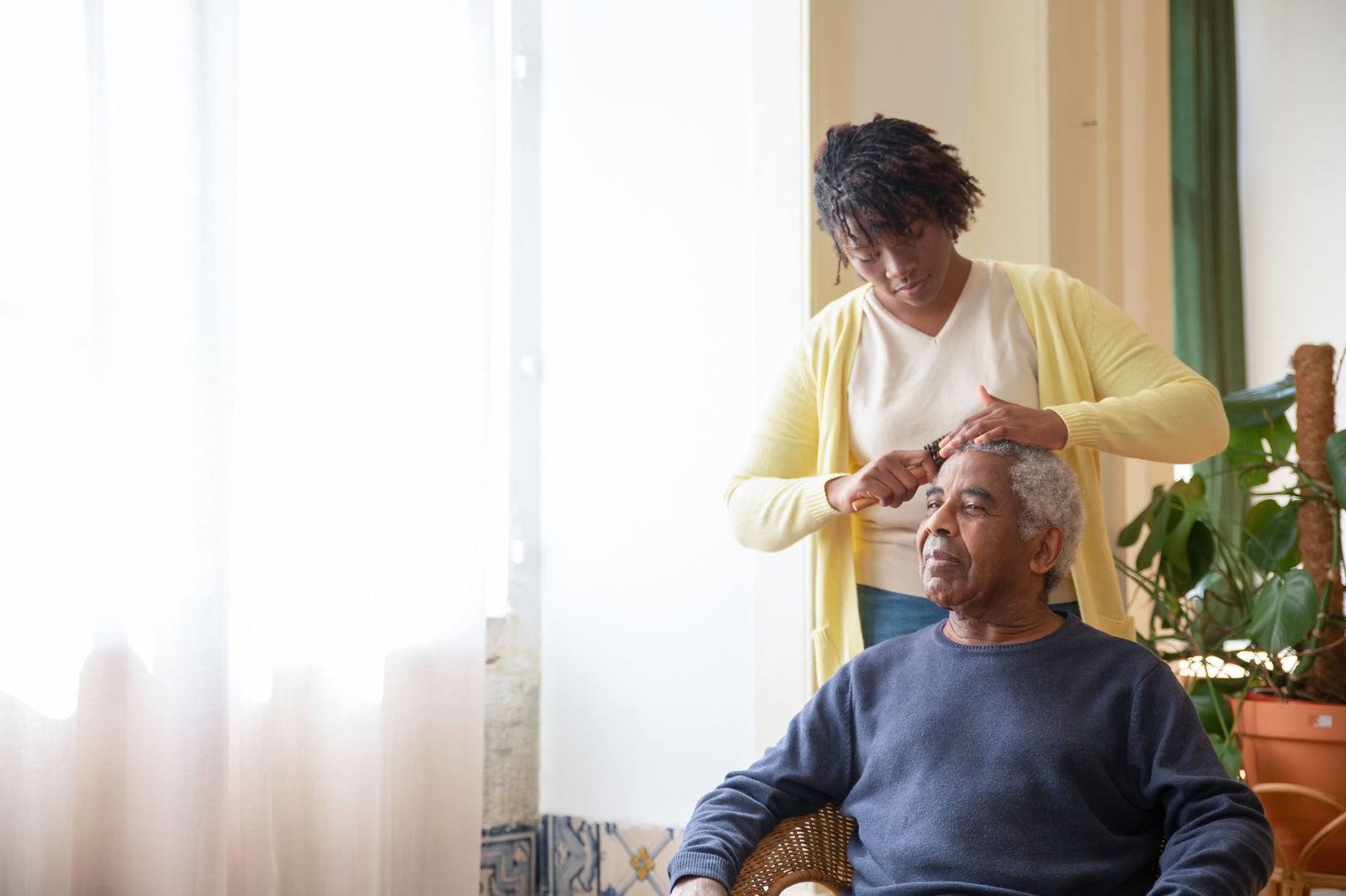 A caregiver gently combing an older man’s hair in a bright, sunlit living room